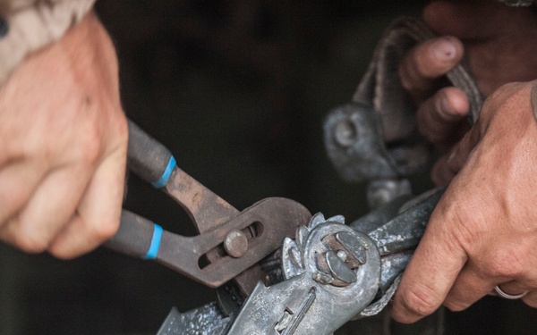 Two individuals are working together with tools on a mechanical fastening device attached to a strap or webbing. One person is using pliers with blue tape on the handle to manipulate a metal buckle or ratchet mechanism. The scene appears to be a close-up of a hands-on mechanical or rigging task.