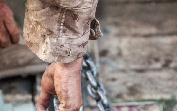 A person wearing desert camouflage military uniform is handling heavy mechanical equipment, gripping a large threaded cylindrical component with a metal chain attached. The hands are visibly dirty and calloused, suggesting physical labor in a field or maintenance environment. The equipment appears to be military or industrial machinery, possibly artillery or vehicle-related hardware.