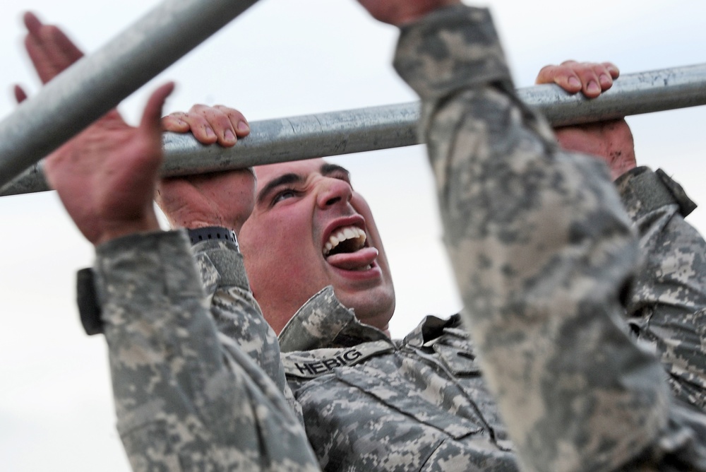 A soldier in US Army ACU (Army Combat Uniform) camouflage is performing pull-ups on a metal bar, captured from a low angle looking upward. The soldier's name tape reads 'HERIG' and their face shows intense physical exertion with mouth open. The image appears to be taken during a physical fitness test or obstacle course training.