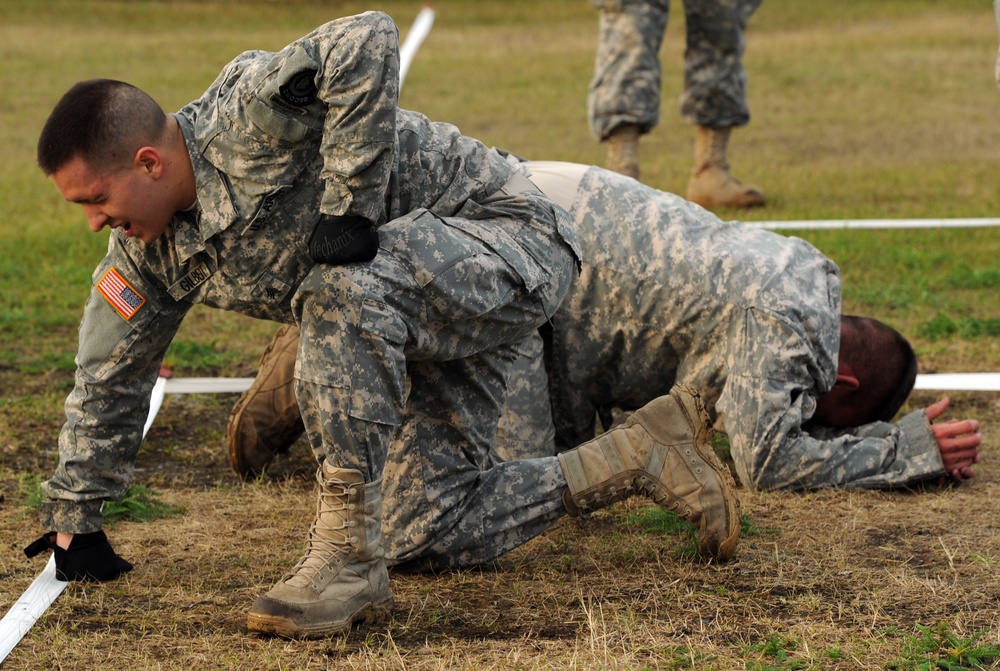 Two U.S. Army soldiers in ACU (Army Combat Uniform) pattern camouflage are engaged in a combatives or ground fighting training exercise on a grass field. One soldier is positioned on top of the other in a dominant ground control position, consistent with military combatives training. A third soldier in uniform is visible standing in the background, likely observing or supervising.