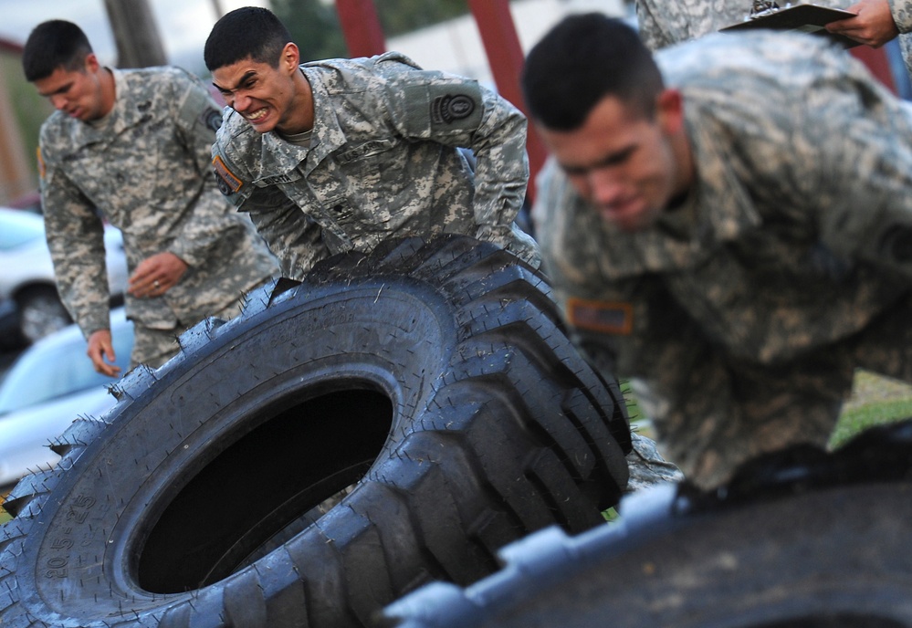Military personnel in US Army ACU (Army Combat Uniform) camouflage are performing a tire-flipping physical training exercise. At least three soldiers are visible actively flipping large heavy-duty tires, with a fourth person in the background appearing to observe or record the activity. The soldiers display visible physical exertion, with strained facial expressions indicating intense effort.