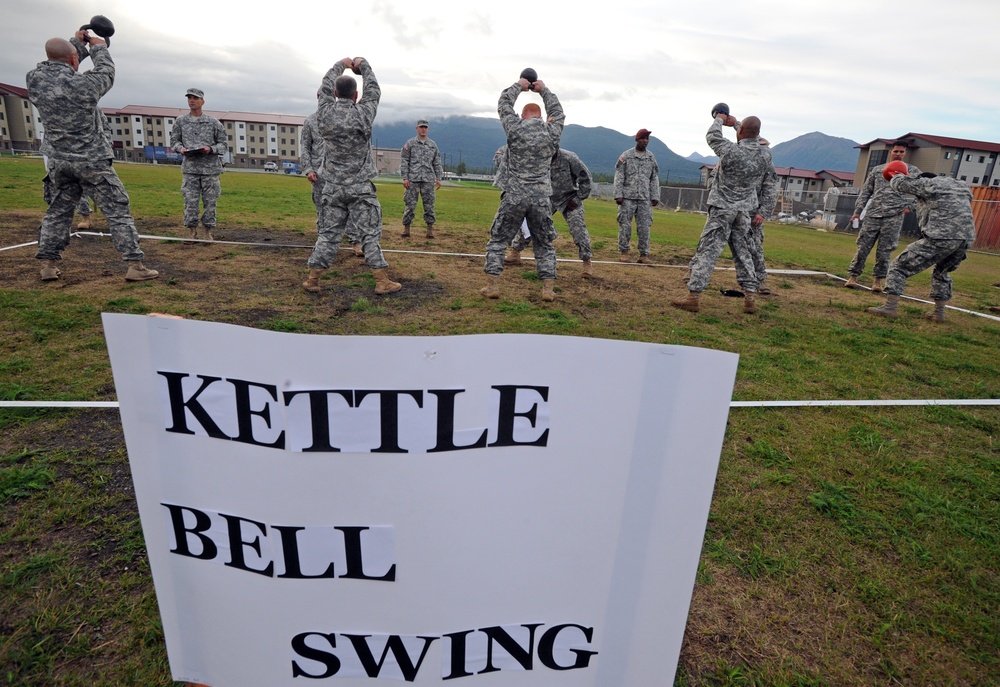 A group of military personnel in camouflage uniforms are performing kettle bell swing exercises on a grassy field at what appears to be a military installation. A large white sign in the foreground reads 'KETTLE BELL SWING', indicating this is a designated fitness station. Mountains and military barracks buildings are visible in the background.