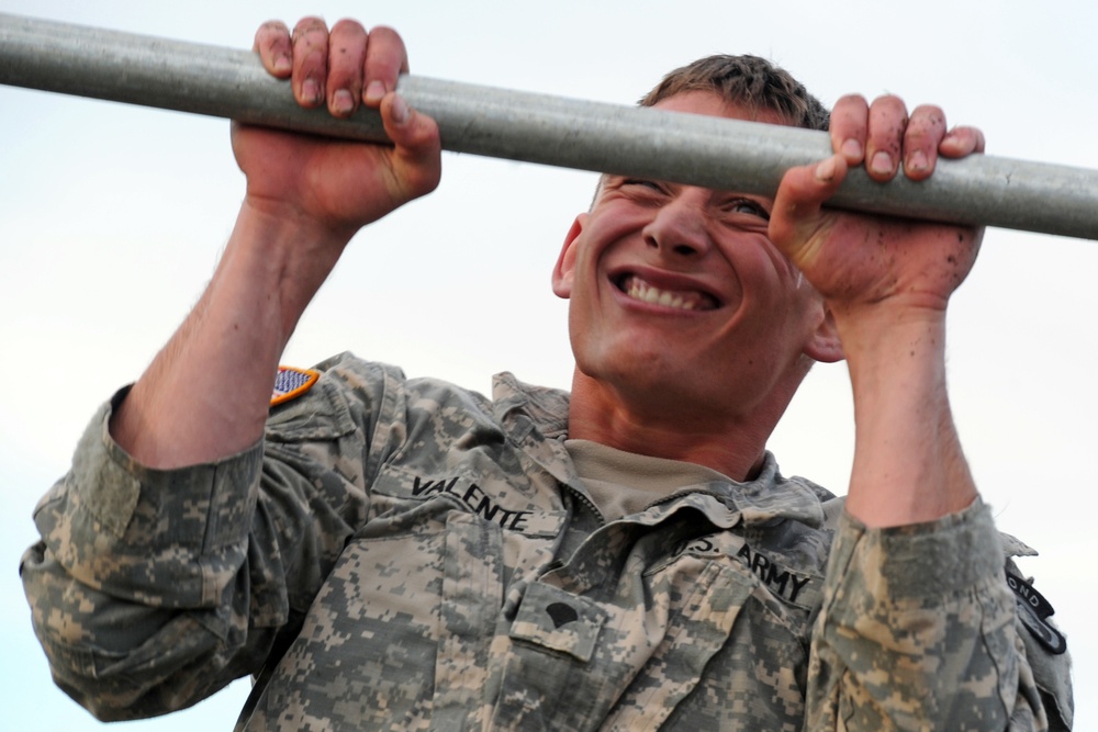 A U.S. Army soldier named Valente is performing pull-ups on a metal bar, wearing ACU (Army Combat Uniform) with digital camouflage pattern. The soldier is smiling broadly despite visible exertion, with dirt/mud visible on hands and face suggesting a physical training or obstacle course event. The photo is taken from a low angle looking up, emphasizing the effort and determination of the subject.