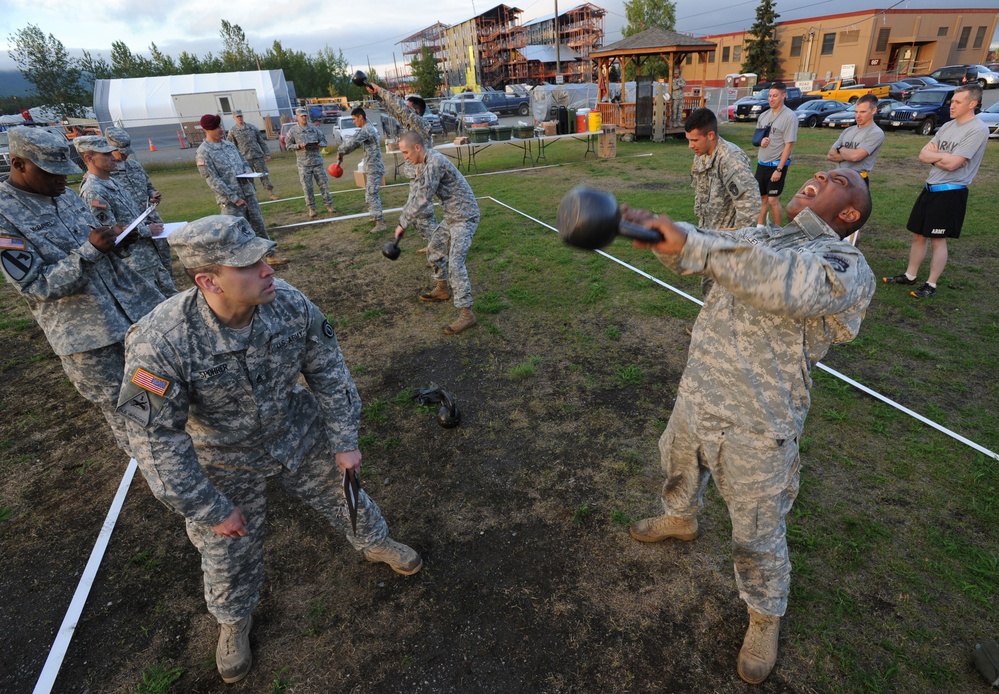A group of U.S. Army soldiers are participating in an outdoor kettlebell training exercise on a grassy field. Multiple soldiers in ACU (Army Combat Uniform) camouflage are performing kettlebell swings and lifts within a roped-off area, while others observe and take notes. The setting appears to be a military installation with buildings, vehicles, and trees visible in the background.