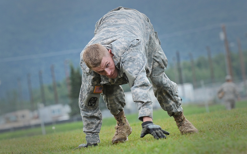 A U.S. Army soldier in ACU (Army Combat Uniform) digital camouflage is performing a physical training exercise on a grass field, appearing to be in a bear crawl or agility drill position. The soldier wears an American flag patch and unit patch on the left sleeve, along with tactical gloves and tan combat boots. The background shows a military installation with utility poles and mountainous terrain.