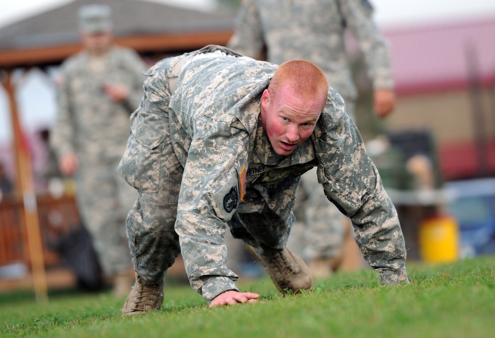 A U.S. Army soldier in ACU (Army Combat Uniform) digital camouflage is performing a low crawl or bear crawl exercise on a grass field. The soldier appears physically exerted, with a flushed face, suggesting intense physical training. Additional soldiers in uniform are visible in the blurred background, indicating a group training or fitness evaluation event.