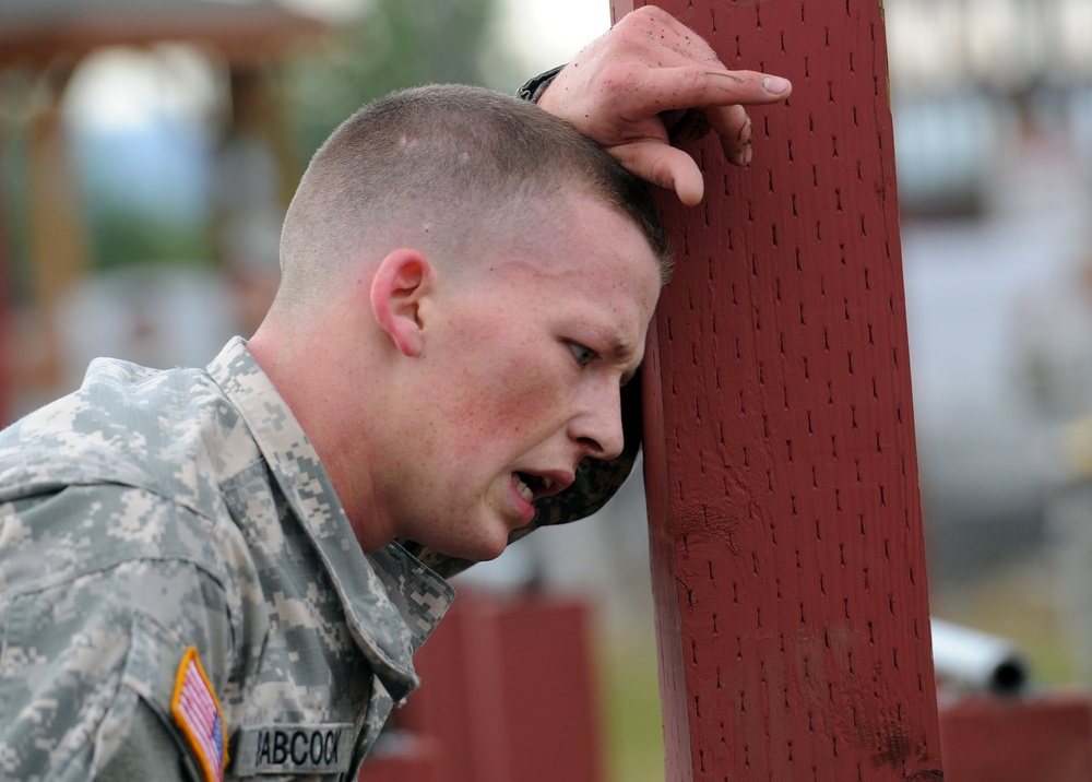 A U.S. Army soldier in ACU (Army Combat Uniform) is leaning his head and hand against a red wooden post, appearing exhausted or in distress. His name tape reads 'BABCOCK' and he wears a U.S. flag patch on his left shoulder. The image captures a moment of physical fatigue, likely during a training exercise or physical fitness event.