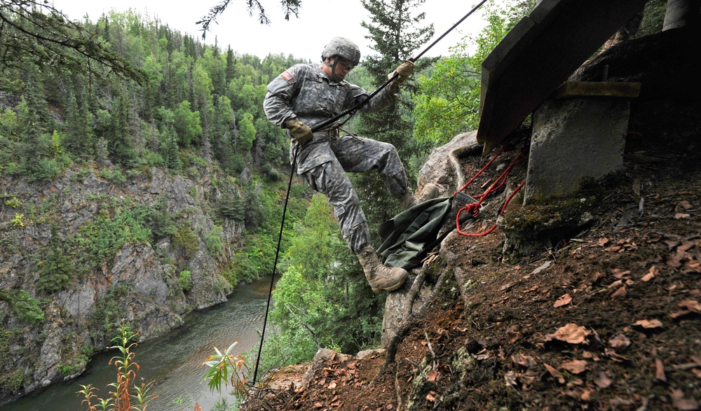 A U.S. Army soldier in full ACU (Army Combat Uniform) is rappelling down a steep cliff face near a river gorge in a forested, mountainous environment. The soldier is gripping a black rappelling rope and appears to be in the process of descending from a concrete structure or bridge abutment. The setting suggests a military training exercise in a wilderness area.