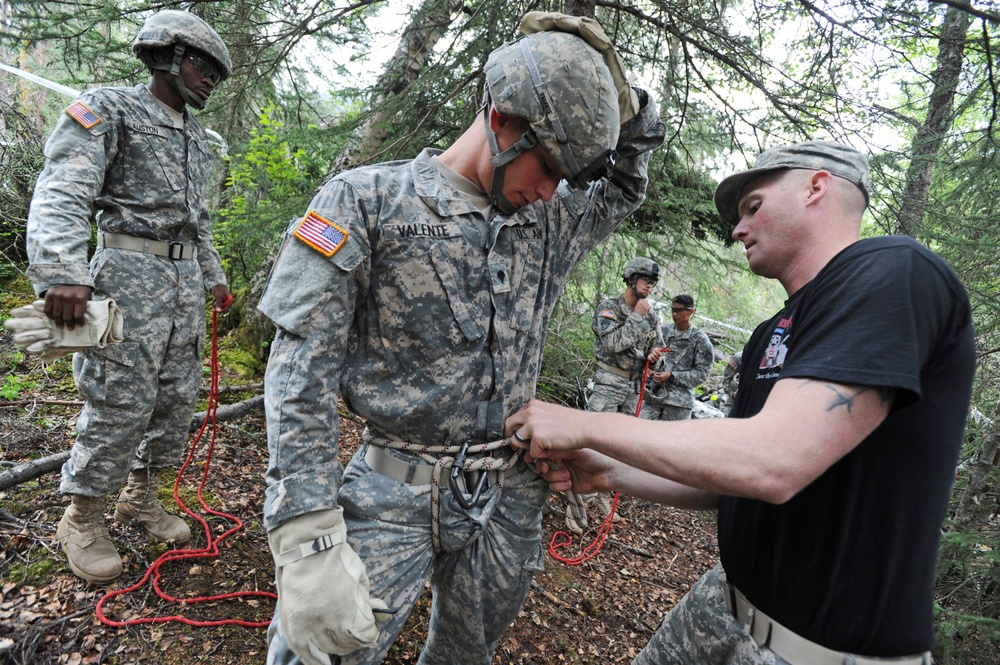 A group of U.S. Army soldiers in ACU (Army Combat Uniform) are engaged in what appears to be rappelling or rope training in a forested area. An instructor in civilian clothing (black t-shirt) is checking and adjusting a harness on a soldier identified by nametape as 'VALENTE'. Additional soldiers are visible in the background observing or awaiting their turn.