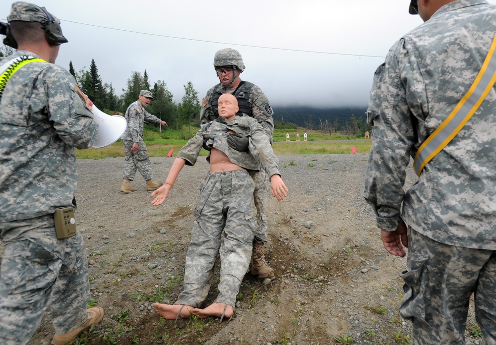 Military personnel are conducting a combat casualty care or rescue training exercise at an outdoor range facility. A soldier in full combat gear is dragging a life-sized training mannequin dressed in military uniform across a gravel surface. Several other soldiers, including an observer with a bullhorn and one wearing a safety vest with reflective stripes, are watching the exercise.