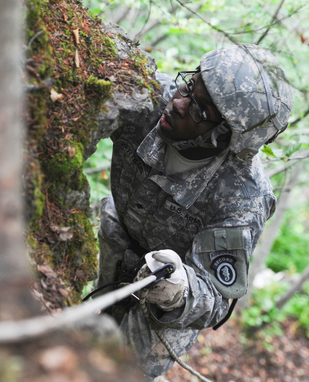 A U.S. Army soldier in ACU (Army Combat Uniform) digital camouflage is rappelling or climbing in a wooded, rocky environment. The soldier is wearing glasses, white gloves, and a helmet, and is gripping a rope with a carabiner attached. Patches on the uniform indicate affiliation with the 2nd Infantry Division Airborne unit.