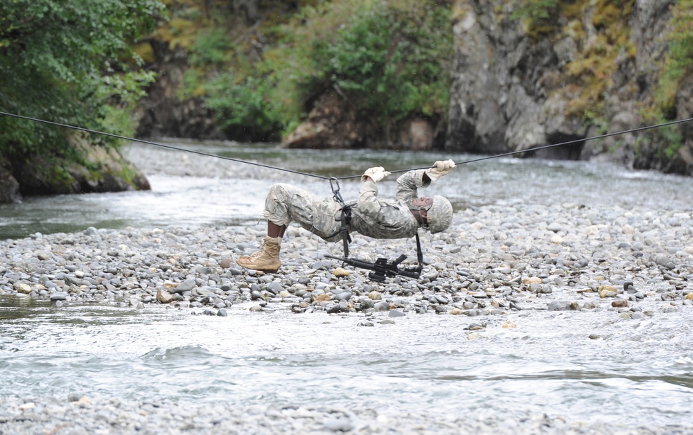 A soldier in camouflage uniform is performing a rope bridge or tyrolean traverse crossing over a rocky riverbed. The individual is suspended horizontally beneath a tensioned rope line, using a pulley or rigging system, with a weapon system also attached and hanging below. The setting appears to be a military training exercise in a mountainous, forested environment.