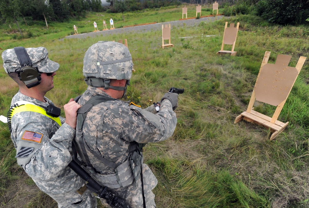Two U.S. Army soldiers are at an outdoor shooting range during what appears to be a training exercise. One soldier wearing a high-visibility yellow vest and hearing protection is observing or supervising while the other soldier in full combat gear aims a pistol at cardboard silhouette targets. Multiple target stands are visible at varying distances across a grassy field.