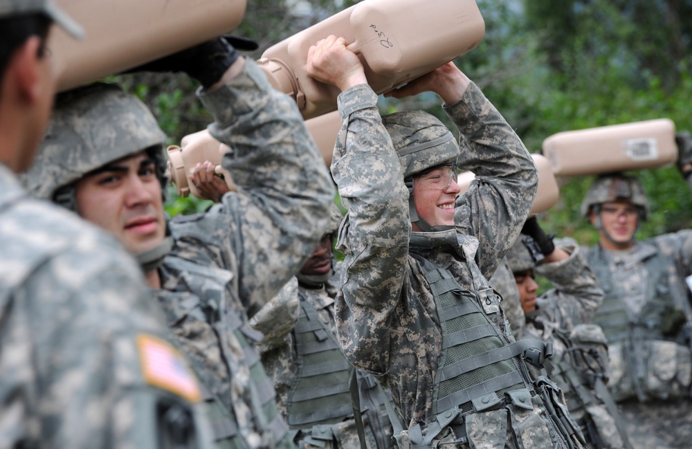 A group of U.S. Army soldiers in ACU (Army Combat Uniform) digital camouflage pattern are participating in what appears to be a physical training or team-building exercise. They are holding large tan/beige containers or equipment cases overhead with both arms extended. One soldier in the center is visibly smiling, suggesting a training or exercise environment rather than combat.