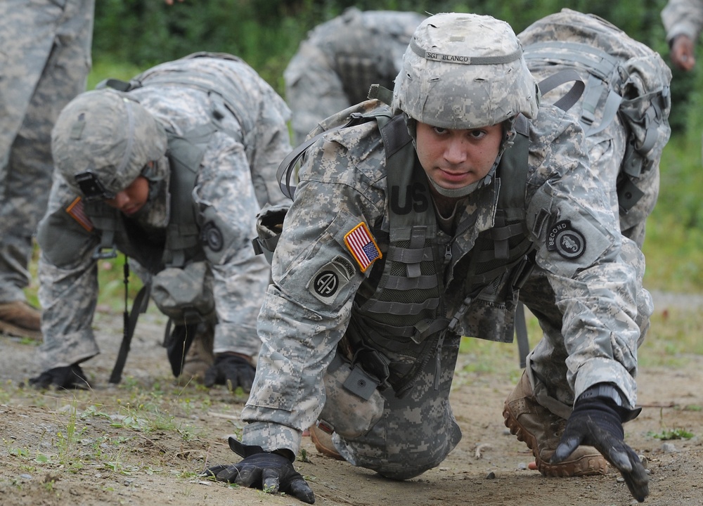 Multiple U.S. Army soldiers in ACU (Army Combat Uniform) digital camouflage are performing a low-crawl exercise on a dirt training course. The foreground soldier, identified by helmet label as 'SGT BLANCO,' wears 82nd Airborne Division patches and is crawling toward the camera with full combat gear. The scene appears to be a military training exercise conducted in an outdoor field environment.