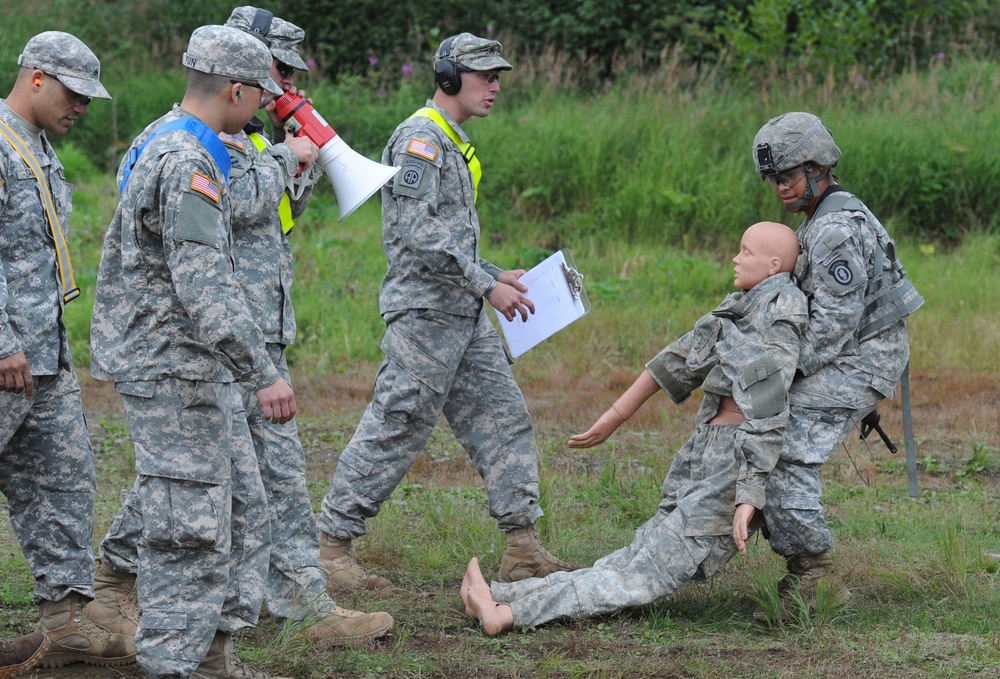 A group of U.S. Army soldiers in ACU (Army Combat Uniform) camouflage are conducting a field training exercise. One soldier is dragging a training mannequin dressed in military uniform, while another soldier with a megaphone and clipboard appears to be directing or evaluating the exercise. The setting is an outdoor grassy field, consistent with a military training area.