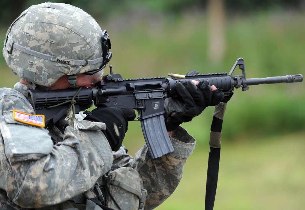 A U.S. Army soldier in ACU (Army Combat Uniform) digital camouflage is aiming an M16/M4-style rifle at a firing range. The soldier is wearing a standard-issue ACH (Advanced Combat Helmet) with the name 'VALENTE' visible on the helmet band. An ejected brass casing is visible mid-air near the ejection port, indicating the weapon has just been fired.