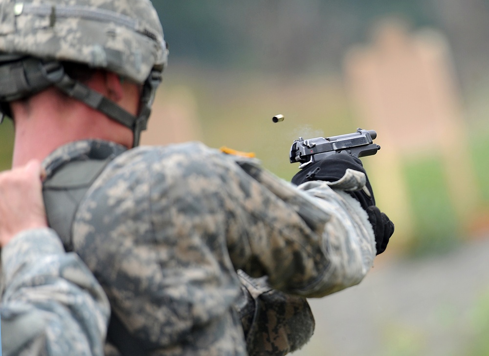 A soldier in U.S. Army ACU (Army Combat Uniform) digital camouflage is firing a semi-automatic pistol at a shooting range. A spent brass casing is visibly ejecting from the pistol's ejection port, captured mid-air by high-speed photography. Silhouette targets are visible in the blurred background.