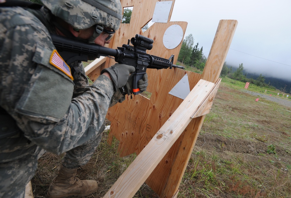 A U.S. Army soldier in ACU (Army Combat Uniform) camouflage is conducting a weapons training exercise at an outdoor shooting range. The soldier is aiming an M4 carbine rifle around a plywood barricade toward downrange targets. The setting appears to be a military training facility, likely in Alaska based on the forested, mountainous background.