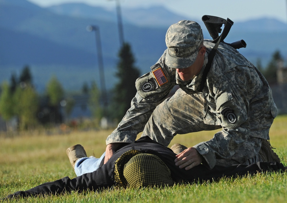 A U.S. Army soldier in ACU (Army Combat Uniform) is kneeling over a person lying on a grass field, appearing to perform a tactical casualty care or combat lifesaver drill. The soldier wears a U.S. flag patch and unit patch on the left sleeve, and has a rifle slung across their back. The setting appears to be a military training area with mountains and trees visible in the background.
