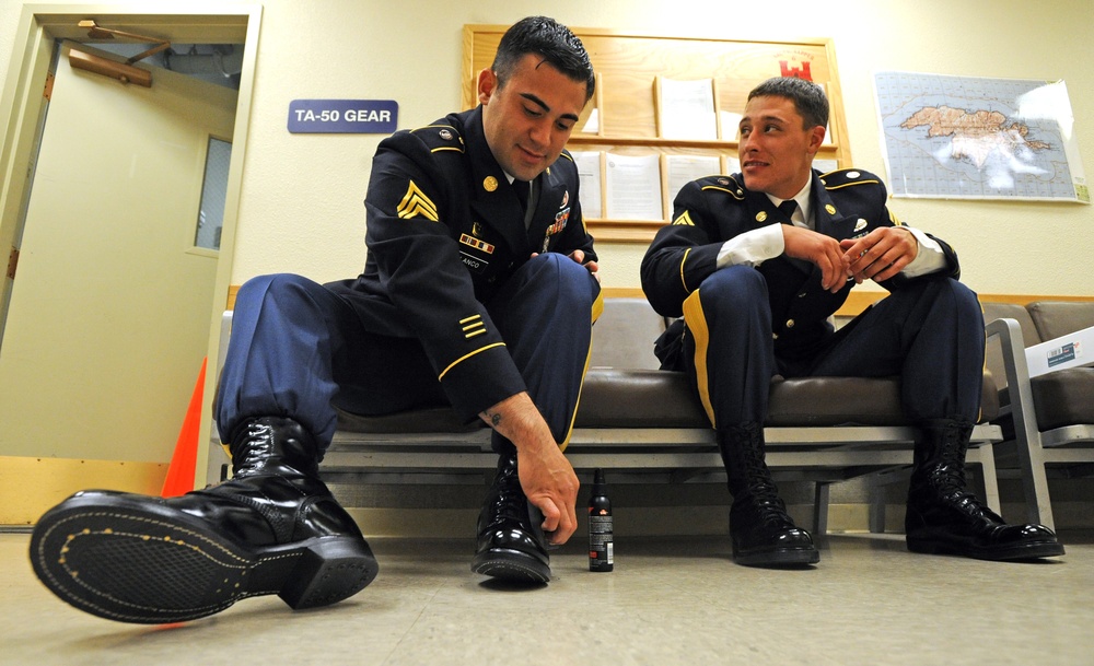 Two U.S. Army soldiers in dress blue uniforms are seated on a bench in what appears to be a military administrative area. The soldier on the left, bearing sergeant chevrons, is polishing or inspecting his black dress boots, while the soldier on the right watches and converses. A sign reading 'TA-50 GEAR' is visible on the wall behind them.