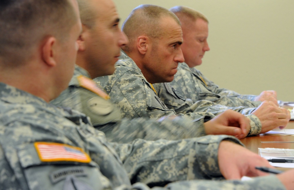 Several U.S. Army soldiers in ACU (Army Combat Uniform) digital camouflage are seated at a table, appearing to attend a briefing or meeting. The soldiers are attentive and facing the same direction, with some holding pens and papers. The setting appears to be an indoor military conference or classroom environment.