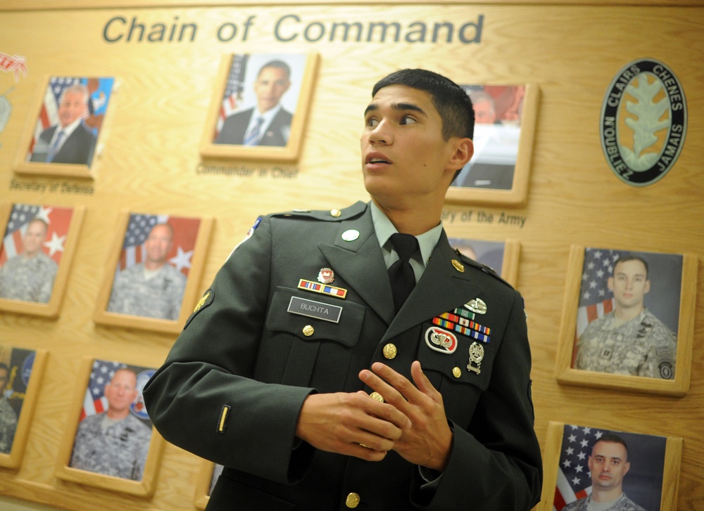 A U.S. Army soldier in Class A dress green uniform stands in front of a 'Chain of Command' display board. The soldier, identified by nameplate as 'BUCHTA', appears to be speaking or presenting, with hands gesturing. The wall behind features framed photographs of military and civilian leadership in the chain of command.