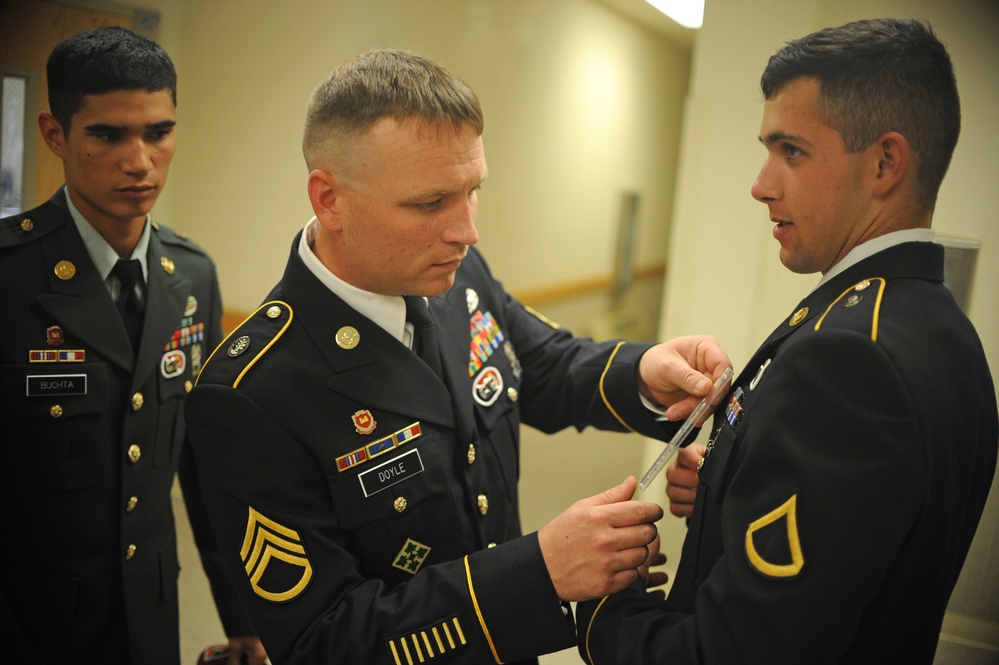 Three U.S. Army soldiers in dress blue uniforms are gathered in what appears to be an indoor corridor or hallway. A Staff Sergeant named Doyle is using a ruler or measuring tool to check the precise placement of ribbons or insignia on a Private First Class's uniform. A third soldier named Buchta observes in the background.