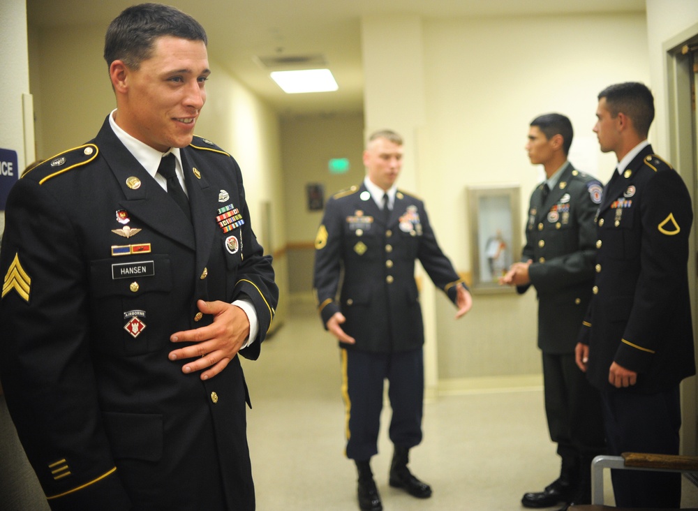 Four U.S. Army soldiers are gathered in an indoor hallway wearing Army Service Uniforms (dress blues). The soldier in the foreground is identified by his name tape as 'HANSEN' and wears the rank of Staff Sergeant with an Airborne tab and multiple ribbons and badges. The other soldiers in the background appear to be conversing or waiting in what looks like a military building corridor.