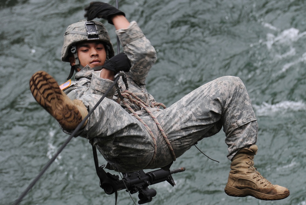 A U.S. Army soldier in full combat uniform is suspended mid-air on a rope or cable system over water, performing what appears to be a rappelling or rope-crossing exercise. The soldier is wearing ACU (Army Combat Uniform) digital camouflage, a combat helmet, black gloves, and tan combat boots, with a harness and rope system securing them. A pair of binoculars is visible attached to their gear.