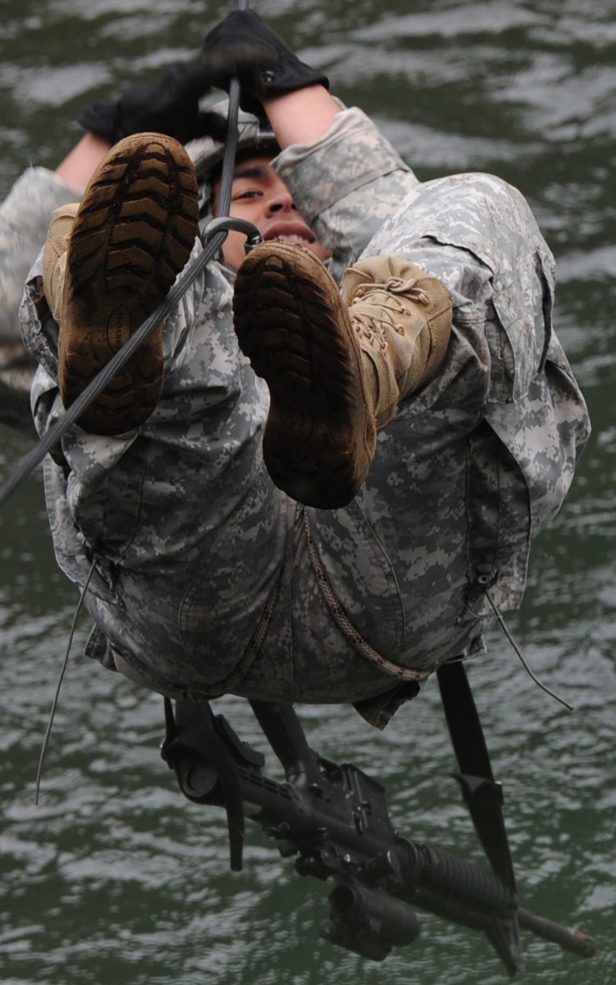 A soldier in US Army ACU (Army Combat Uniform) digital camouflage is traversing a rope or cable line over a body of water, suspended horizontally with feet raised. The soldier is wearing tan combat boots and black gloves, carrying a rifle and a large rucksack. The individual appears to be smiling, suggesting a training exercise rather than a combat situation.