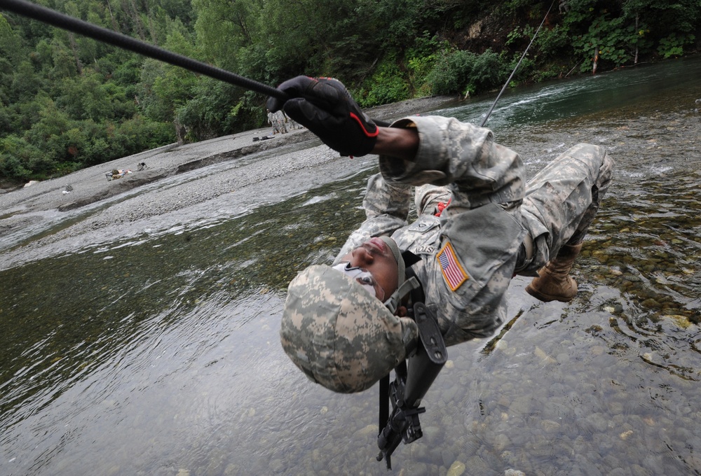 A U.S. Army soldier in ACU (Army Combat Uniform) is traversing a rope line over a rocky river, suspended upside down or at an extreme angle. The soldier is gripping a rope with gloved hands while using a rappelling/rigging device, with a river and forested rocky terrain visible below and in the background. This appears to be a military training exercise involving rope bridge or water crossing techniques.