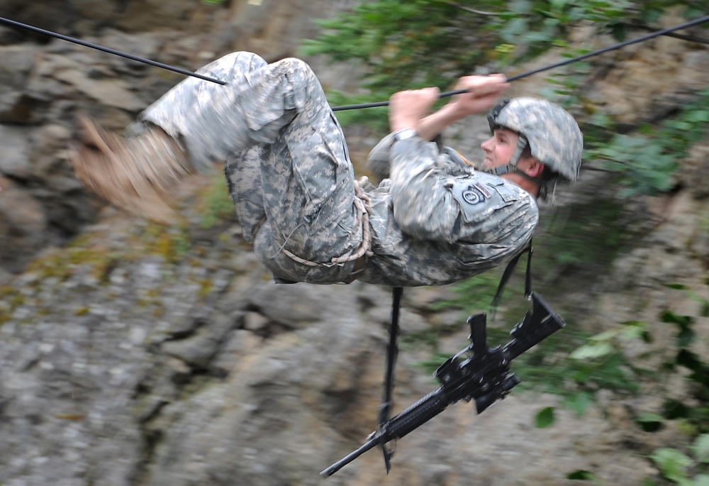 A soldier in US Army ACU (Army Combat Uniform) digital camouflage is traversing a rope or cable line, suspended upside-down or at an extreme angle above rocky terrain. The soldier is wearing a combat helmet and carrying a rifle slung beneath them. The motion blur in the image suggests rapid movement along the line, consistent with a military training exercise.