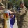 81st Civil Affairs Battalion welcome home ceremony Two U.S. Army soldiers in MultiCam/OCP camouflage uniforms are handling a purple ceremonial guidon or unit colors adorned with an eagle emblem and multiple streamer ribbons. The ceremony appears to be a formal military colors event, likely a casing or uncasing of colors, activation, or inactivation ceremony. Additional soldiers in formation are visible in the background near a brick building.