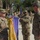 81st Civil Affairs Battalion welcome home ceremony A U.S. Army military ceremony is taking place outdoors, likely a casing or uncasing of colors or an award streamer presentation. Three soldiers in the foreground are handling a purple unit guidon/flag with gold and green streamers, while formations of soldiers stand at attention in the background. The setting appears to be a military installation with brick buildings and trees visible.