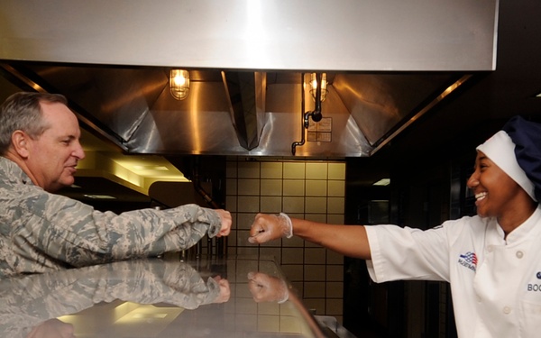 A military service member in camouflage uniform is interacting with a food service worker in a military dining facility (DFAC). The cook, wearing a white chef's coat with the name tag 'BOOKER' and a blue chef's hat, is smiling and extending a fist bump toward the uniformed individual across a stainless steel serving counter. The setting appears to be an active military base cafeteria kitchen.