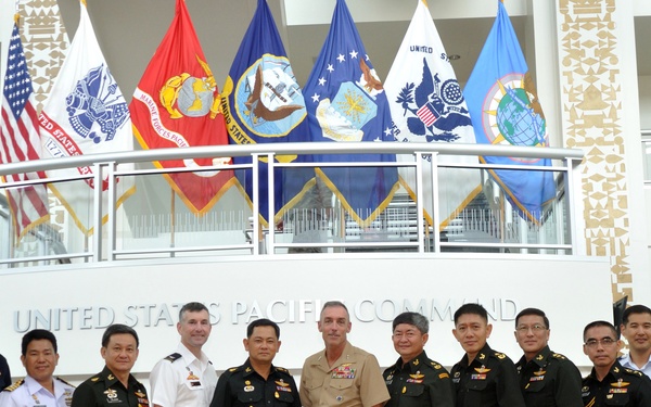 A group of approximately 14 military officers and civilians pose for a formal group photograph inside the United States Pacific Command (USPACOM) headquarters. The group includes personnel in various military uniforms representing multiple nations and branches of service, along with two civilians in business attire. Multiple U.S. military branch flags are displayed on an upper balcony behind the group.