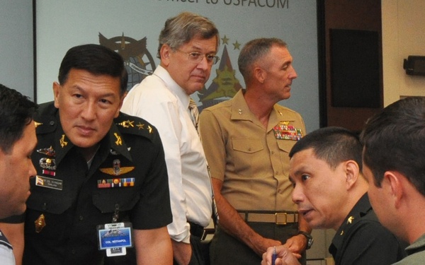 A multinational military meeting or briefing is taking place in a conference room, with officers from multiple nations engaged in discussion around a table. The presentation screen in the background references 'USPACOM' (United States Pacific Command) and displays a Philippine military emblem, suggesting a joint military coordination event. Personnel in various national uniforms are conversing, with documents and binders visible on the table.