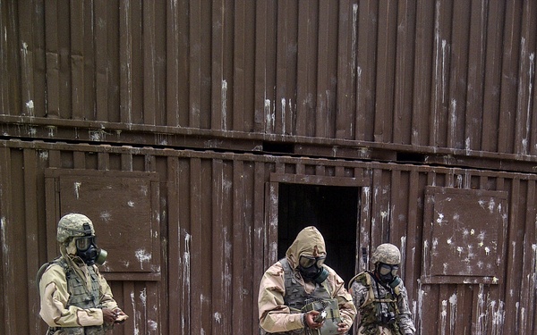 Three military personnel in full CBRN (Chemical, Biological, Radiological, Nuclear) protective gear are conducting what appears to be a training exercise outside a weathered brown metal building. All individuals are wearing desert three-color camouflage uniforms, tactical vests, and military-issue gas masks. One person is exiting through a low doorway of the structure while another holds a piece of equipment, suggesting a structured drill or evaluation scenario.