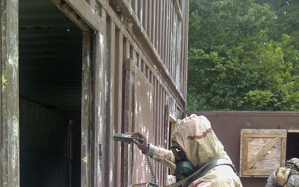 A military personnel wearing full MOPP (Mission Oriented Protective Posture) gear, including a gas mask and desert camouflage CBRN protective suit, is using a detection instrument near the wall of a corrugated metal structure. A second similarly equipped soldier is visible in the background near wooden crates. This appears to be a CBRN (Chemical, Biological, Radiological, Nuclear) training exercise at a military training facility.