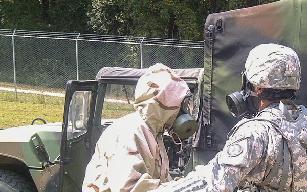 Two military personnel wearing gas masks are conducting what appears to be a CBRN (Chemical, Biological, Radiological, Nuclear) training exercise near a military HMMWV (Humvee). One soldier is wearing a desert camouflage MOPP (Mission Oriented Protective Posture) suit, while the other is in ACU (Army Combat Uniform) pattern. The scene takes place outdoors near a chain-link fence with trees in the background.