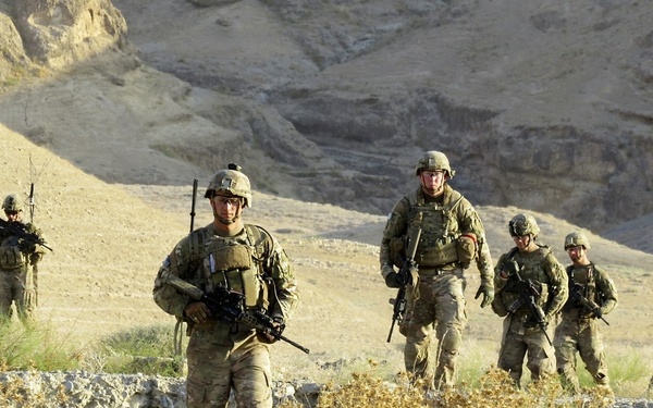 A group of approximately seven soldiers in full combat gear are conducting a patrol through an arid, rocky terrain with sparse vegetation. They are wearing camouflage uniforms and helmets, carrying assault rifles and tactical equipment. The landscape features eroded sandy hills and cliffs in the background, consistent with a desert or semi-arid environment.