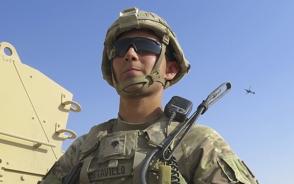 A U.S. Army soldier in full combat gear is photographed from a low angle against a clear blue sky in what appears to be an arid, desert environment. The soldier is wearing Multicam (OCP) camouflage, a combat helmet, ballistic sunglasses, and body armor with a radio and weapon visible. A military vehicle (likely an MRAP or similar armored vehicle) is visible to the left, and a helicopter can be seen in the distant sky.