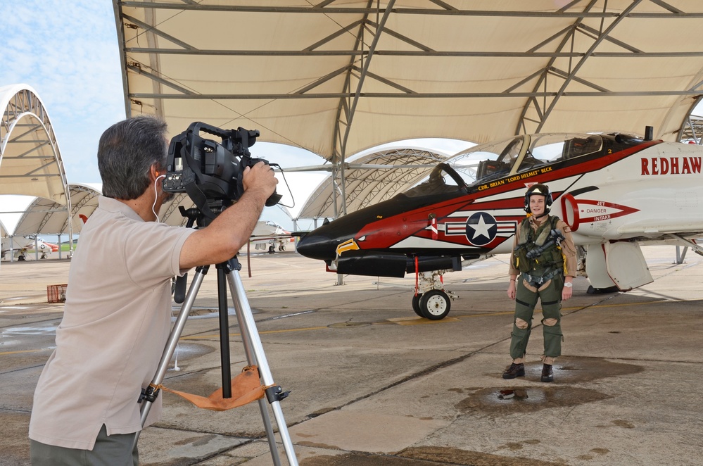 A videographer on a tripod films a person in flight gear standing beside a distinctively painted military jet aircraft under a hangar canopy. The aircraft features a bold red, black, and white eagle design with the name 'CDR BRIAN LORD HELMET BECK' and the word 'REDHAW...' (likely 'REDHAWKS') visible on the fuselage. The setting appears to be a military air base flight line.