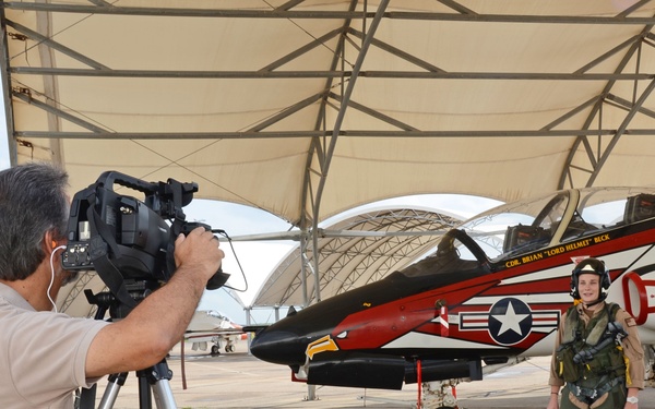 A videographer on a tripod films a person in flight gear standing beside a distinctively painted military jet aircraft under a hangar canopy. The aircraft features a bold red, black, and white eagle design with the name 'CDR BRIAN LORD HELMET BECK' and the word 'REDHAW...' (likely 'REDHAWKS') visible on the fuselage. The setting appears to be a military air base flight line.