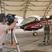 A videographer on a tripod films a person in flight gear standing beside a distinctively painted military jet aircraft under a hangar canopy. The aircraft features a bold red, black, and white eagle design with the name 'CDR BRIAN LORD HELMET BECK' and the word 'REDHAW...' (likely 'REDHAWKS') visible on the fuselage. The setting appears to be a military air base flight line.