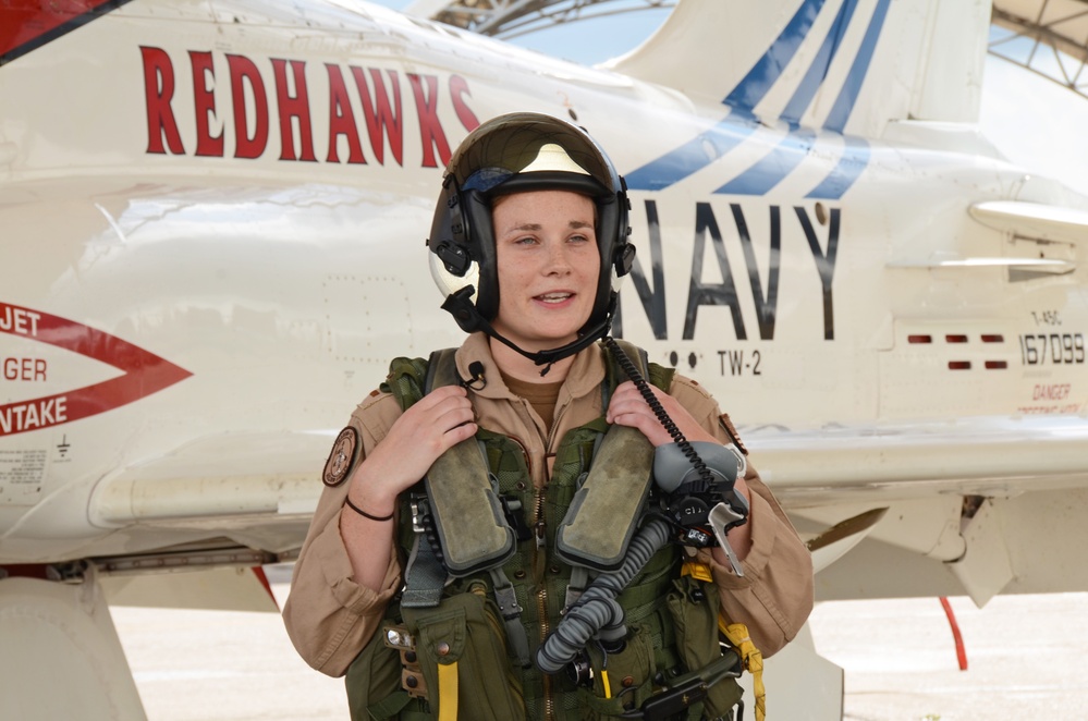 A naval aviator in full flight gear stands in front of a U.S. Navy T-45C Goshawk jet trainer aircraft. The aircraft belongs to Training Wing Two (TW-2), known as the 'Redhawks,' with tail number 167099. The pilot is wearing a flight helmet, survival vest, and tan flight suit, suggesting this may be a pre- or post-flight photo.