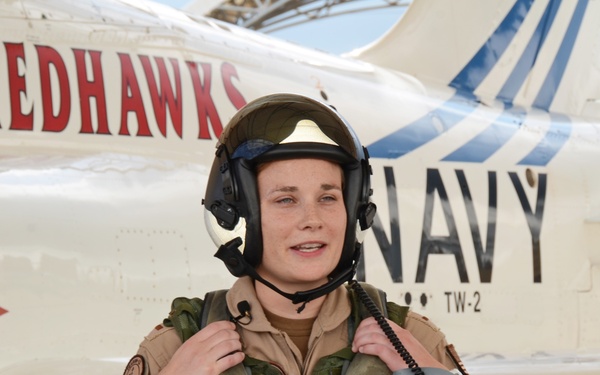 A naval aviator in full flight gear stands in front of a U.S. Navy T-45C Goshawk jet trainer aircraft. The aircraft belongs to Training Wing Two (TW-2), known as the 'Redhawks,' with tail number 167099. The pilot is wearing a flight helmet, survival vest, and tan flight suit, suggesting this may be a pre- or post-flight photo.