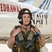 A naval aviator in full flight gear stands in front of a U.S. Navy T-45C Goshawk jet trainer aircraft. The aircraft belongs to Training Wing Two (TW-2), known as the 'Redhawks,' with tail number 167099. The pilot is wearing a flight helmet, survival vest, and tan flight suit, suggesting this may be a pre- or post-flight photo.