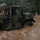 A military camouflage-painted heavy truck is driving through deep, fast-moving floodwaters on what appears to be a submerged road. The brown, turbulent water reaches well up the truck's wheels and undercarriage, suggesting significant flood conditions. At least one uniformed personnel is visible in the cab, indicating an active flood response or rescue operation.